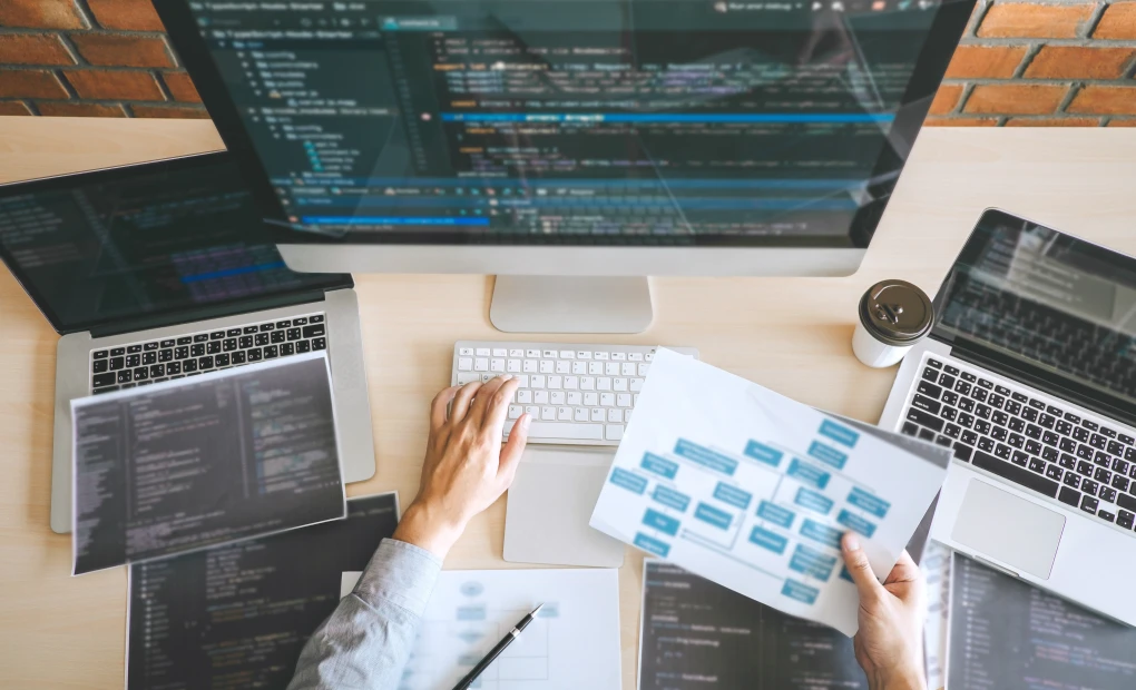 worker sitting in front of two laptops and monitor with code on them